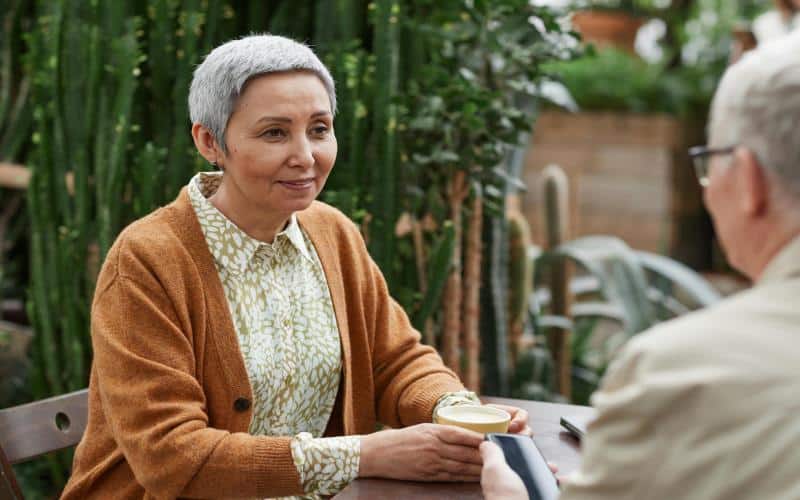 Smiling senior woman enjoying coffee and conversation with a friend in a lush indoor garden café setting.
