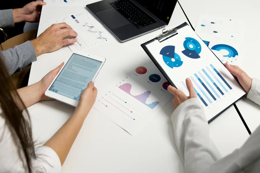 Hands of team members analyzing charts and graphs on paper, tablet, and laptop during a digital marketing meeting.